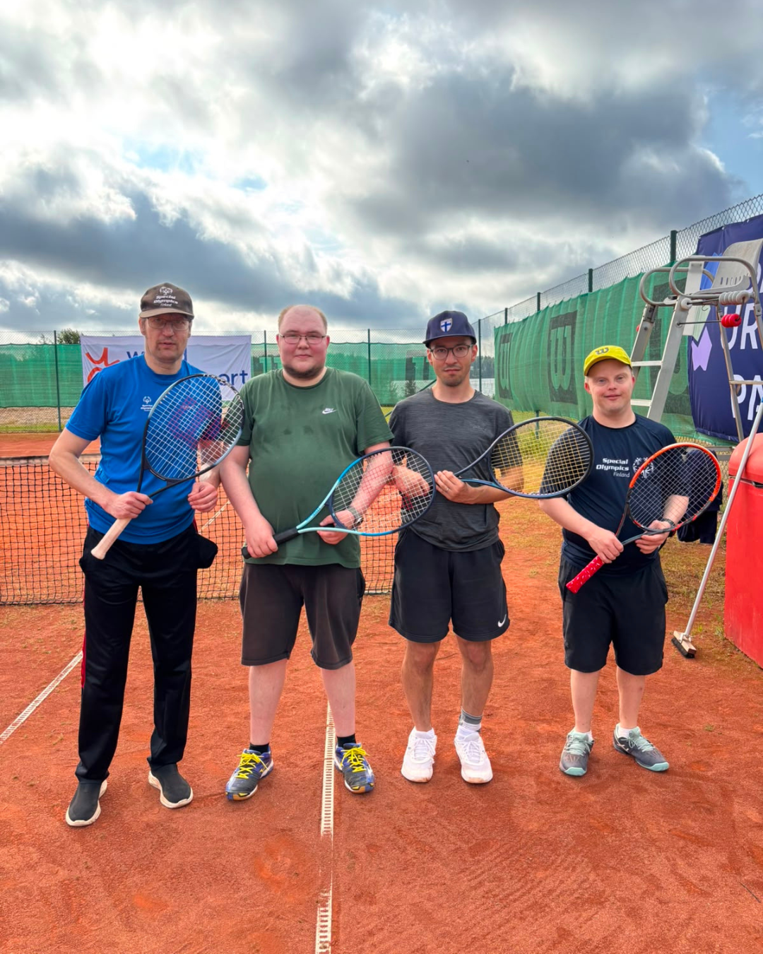 Neljä tennispelaajaa tenniskentällä hymyilemässä kameralle.